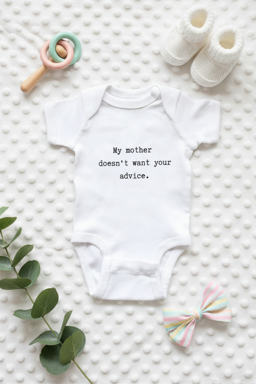 White baby onesie with funny typewriter text surrounded by a baby rattle, tiny shoes, pastel gift bow, and eucalyptus on a soft white surface